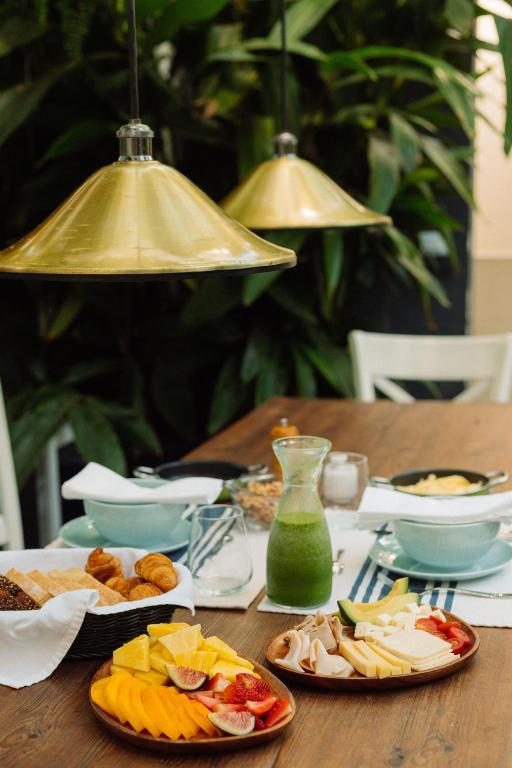 9 Unique Boutique Hotels in Lima 24 A wooden table set for breakfast with plates of assorted sliced fruit, cheese, ham, bread, pastries, and a carafe of green juice—just like the spreads you’d find at boutique hotels in Lima. Gold pendant lights glow above lush green plants in the background.