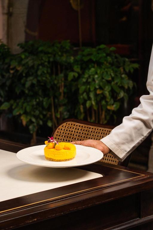 12 BEST Boutique Hotels in Jaipur 50 A person in a white chef’s coat serves a plated yellow dessert topped with fruit and edible flowers on a dark wooden table, capturing the refined elegance often found in boutique hotels in Jaipur, with lush green plants in the background.