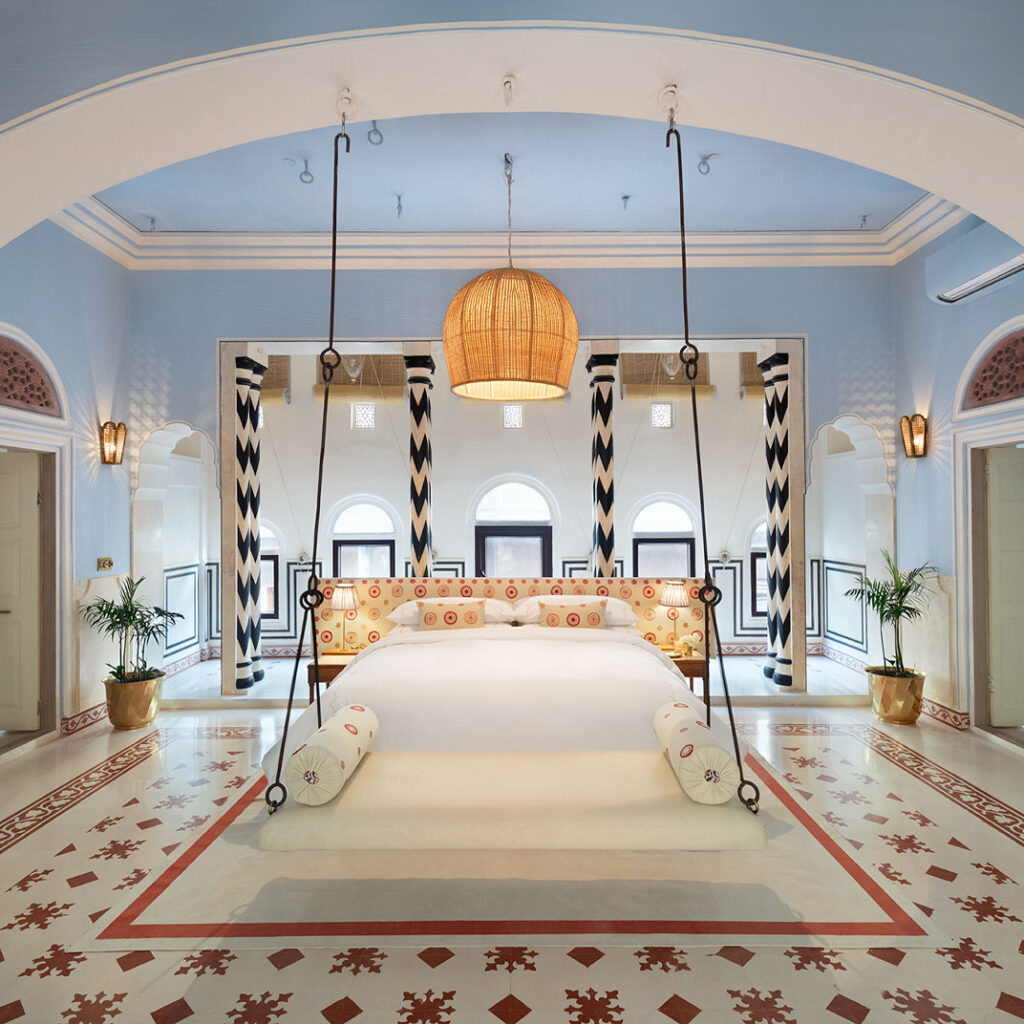 12 BEST Boutique Hotels in Jaipur 37 A spacious, elegant bedroom inspired by boutique hotels in Jaipur features a large white bed suspended by thick ropes, patterned pillows, a woven pendant lamp, arched windows, geometric black and white columns, and decorative tile flooring.