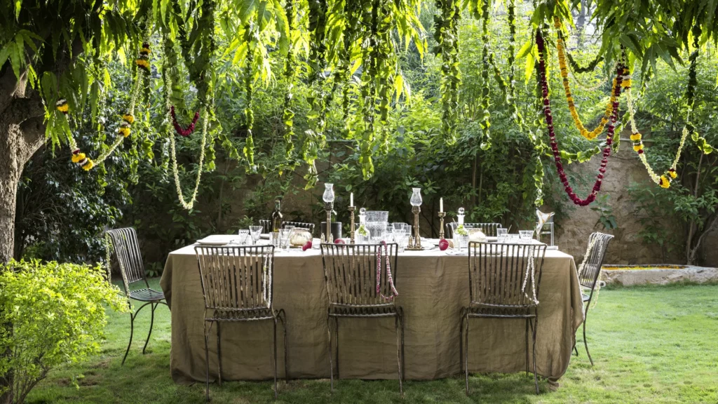 12 BEST Boutique Hotels in Jaipur 5 A garden dining table with a beige tablecloth is set for a meal under hanging greenery and flower garlands, resembling the charm found at boutique hotels in Jaipur, surrounded by metal chairs on a lush green lawn.