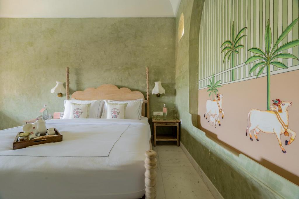 12 BEST Boutique Hotels in Jaipur 27 A cozy bedroom inspired by boutique hotels in Jaipur, featuring a double bed, light green textured walls, wooden headboard, bedside tables, wall lamps, a tea set tray, and a mural with cows and palm trees.