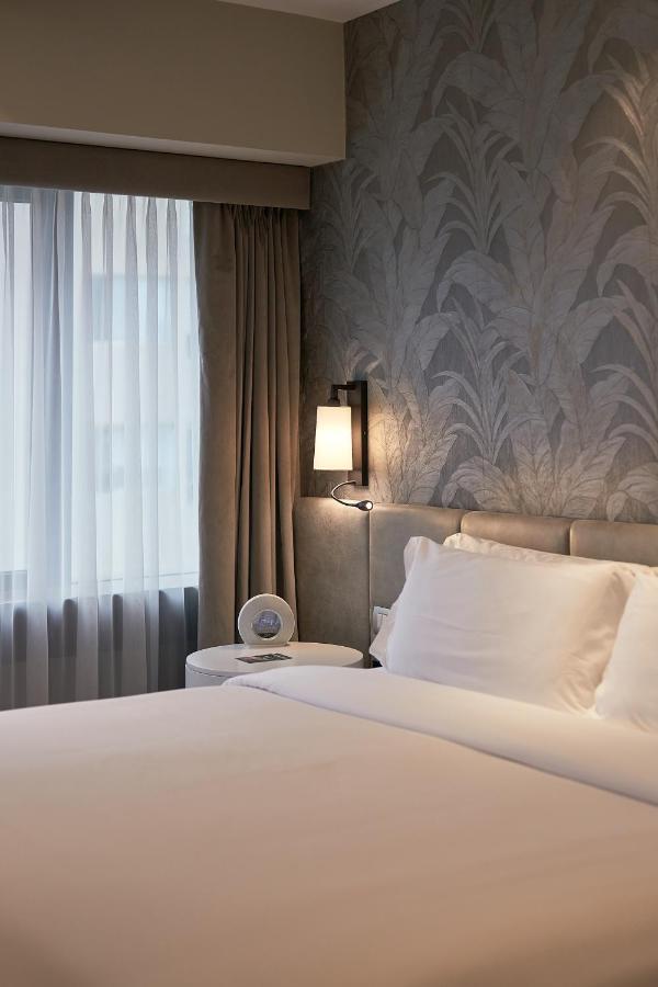 9 Unique Boutique Hotels in Lima 13 A neatly made bed with white linens sits beside a round table, lamp, and alarm clock. Light filters through sheer curtains onto leafy wallpaper, evoking the calm style found in boutique hotels across Lima, Peru.