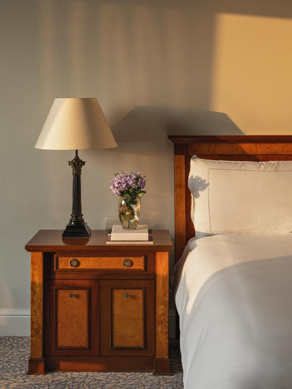 9 Unique Boutique Hotels in Lima 33 A wooden nightstand with a lamp, a vase of purple flowers, and two books sits beside a neatly made bed with white linens, evoking the cozy elegance found in boutique hotels in Lima, all bathed in warm sunlight from the left.