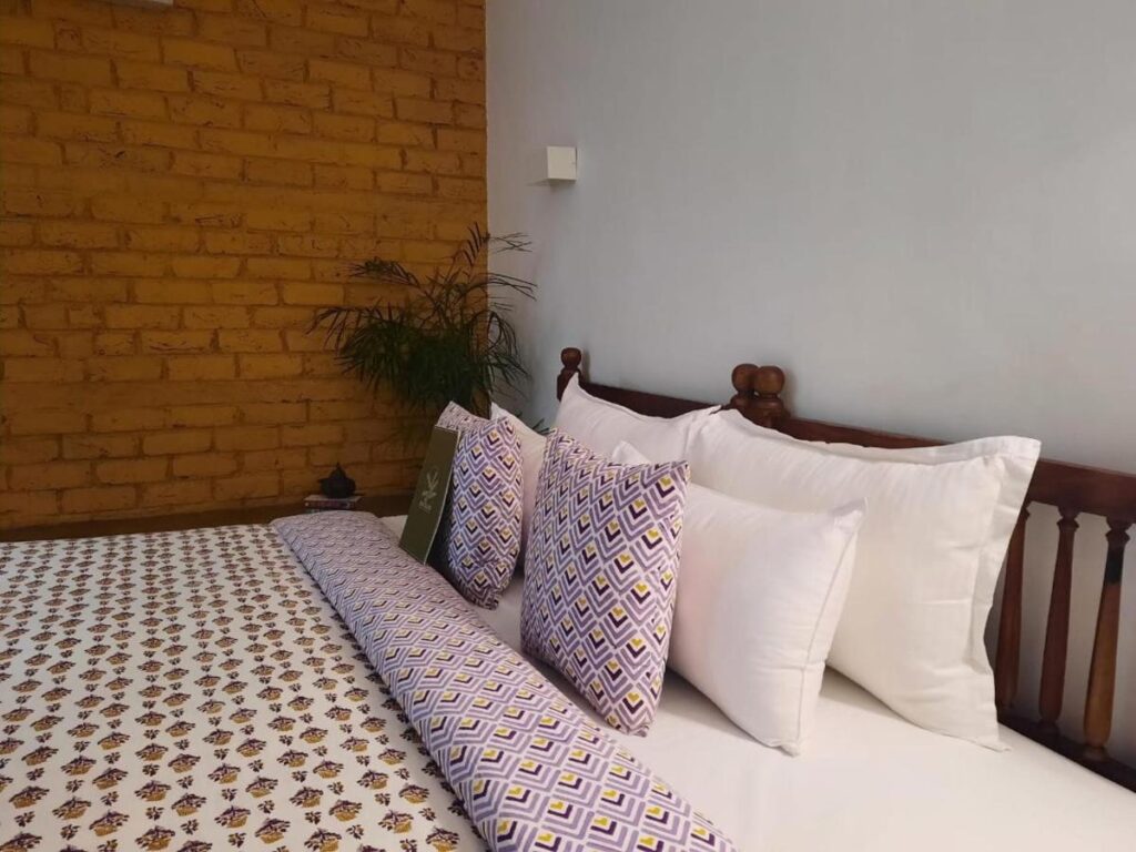 12 BEST Boutique Hotels in Jaipur 19 A neatly made bed with patterned purple and yellow pillows and matching bedspread, set against a wooden headboard beside a yellow brick wall and lush green potted plant, evokes the vibrant charm of boutique hotels in Jaipur.