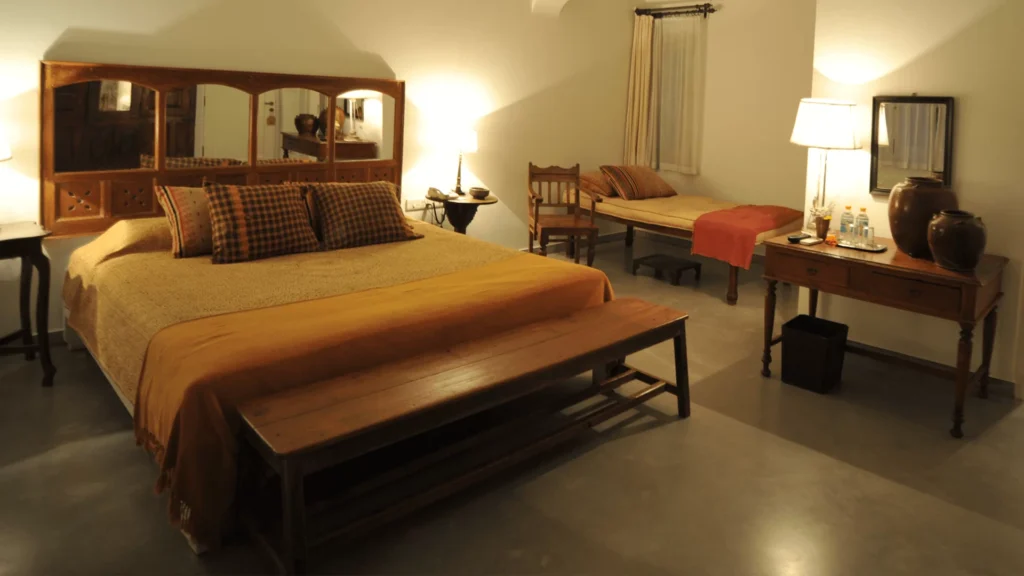 12 BEST Boutique Hotels in Jaipur 6 A cozy bedroom inspired by boutique hotels in Jaipur, with a large bed, decorative pillows, a wooden bench at the foot, single bed by the window, desk with a lamp, bottled water, jars, and chair—all warmly lit.
