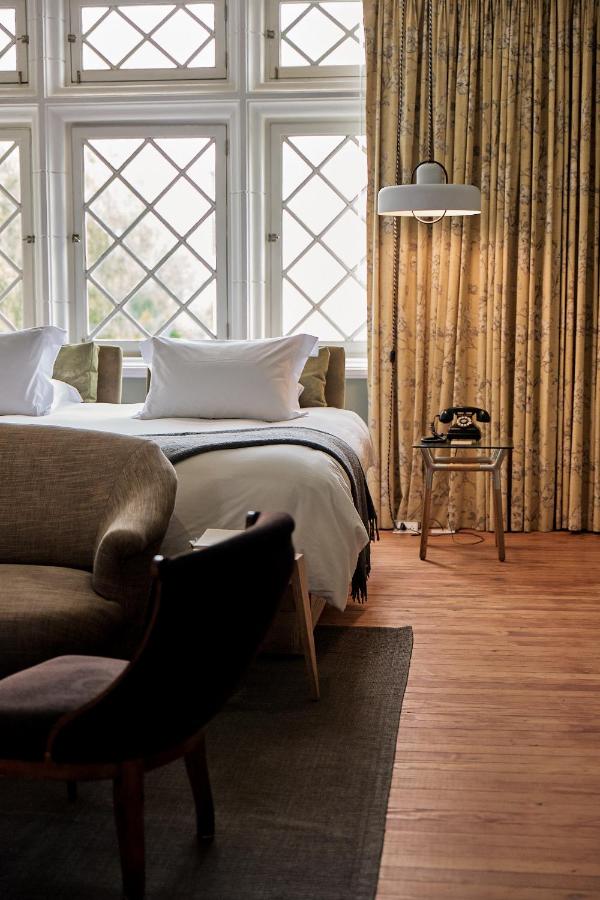 9 Unique Boutique Hotels in Lima 5 A cozy bedroom in one of Lima's boutique hotels, featuring large lattice windows, a double bed with white linens, vintage armchairs, a side table with a black rotary phone, and beige patterned curtains on a wooden floor.