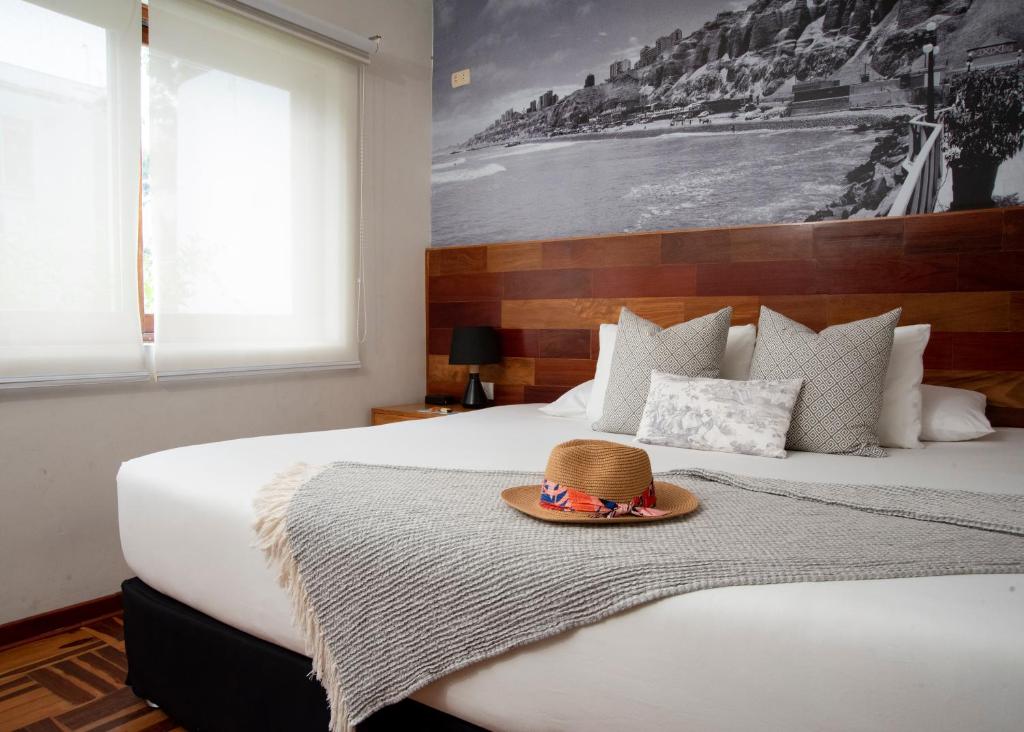 9 Unique Boutique Hotels in Lima 19 A cozy bedroom with a large bed, gray blanket, decorative pillows, and a straw hat on top—reminiscent of the charm found in boutique hotels in Lima. A black-and-white seaside mural graces the wall as soft light pours through white blinds.