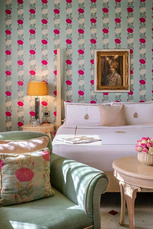 12 BEST Boutique Hotels in Jaipur 12 A cozy bedroom reminiscent of boutique hotels in Jaipur features a green armchair, a round table with pink flowers, a bed with white and beige linens, a lamp, and floral wallpaper. A framed portrait hangs above the bed.