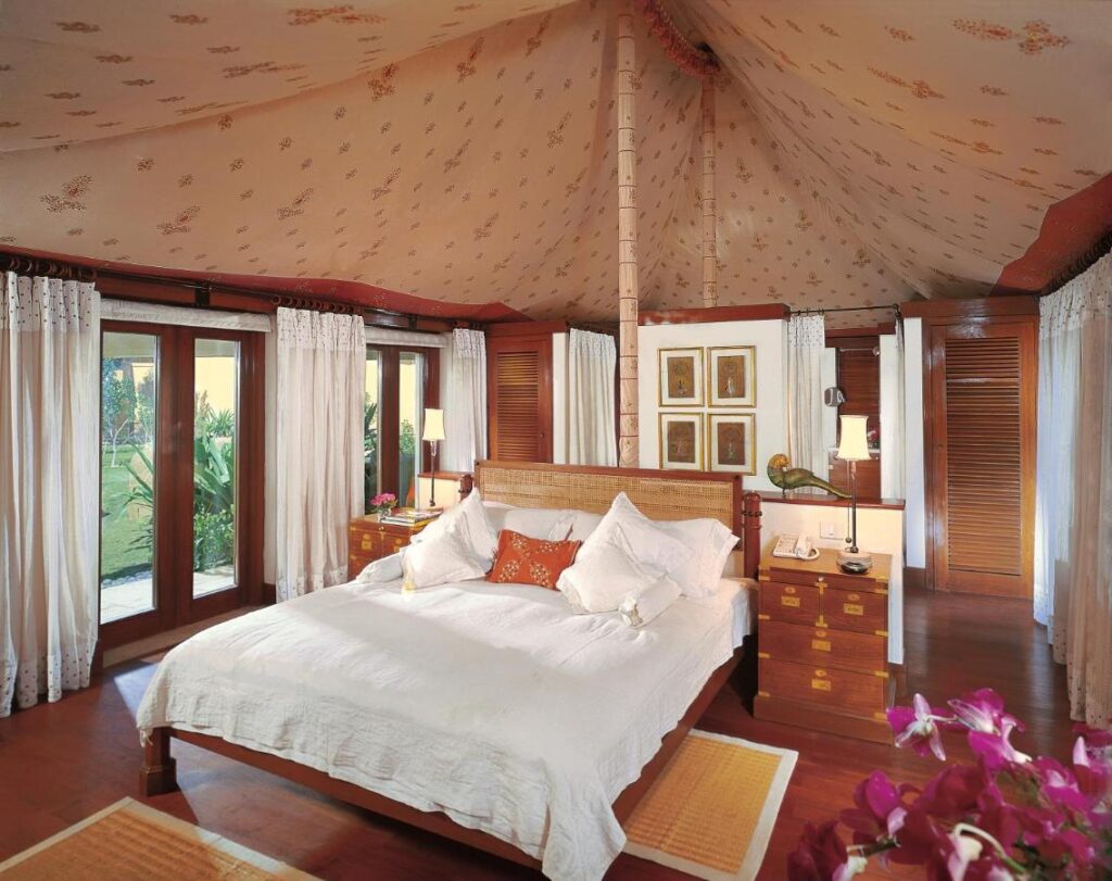 12 BEST Boutique Hotels in Jaipur 45 A spacious, elegant bedroom inspired by boutique hotels in Jaipur, with a high tented ceiling, large windows with sheer curtains, a wooden bed with white bedding, orange accent pillows, bedside tables, lamps, and fresh flowers.