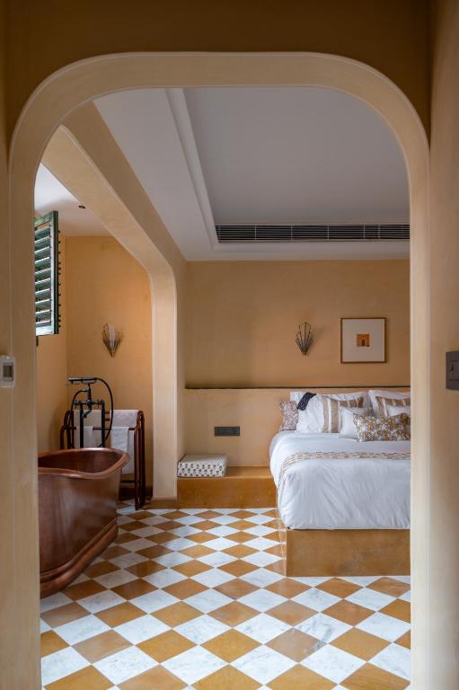 12 BEST Boutique Hotels in Jaipur 20 A stylish bedroom inspired by boutique hotels in Jaipur, with warm beige walls, a wooden bathtub, a white bed with patterned pillows, and a checkered tile floor, viewed through an arched doorway as sunlight streams in through shuttered windows.