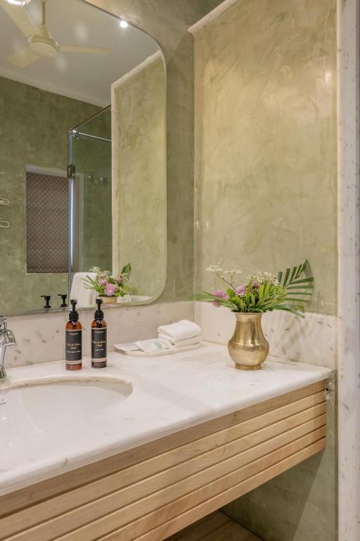 12 BEST Boutique Hotels in Jaipur 26 A modern bathroom inspired by boutique hotels in Jaipur, featuring green marble walls, a large mirror, a white countertop with wooden accents, two bottles of toiletries, neatly folded towels, and a brass vase with fresh flowers.