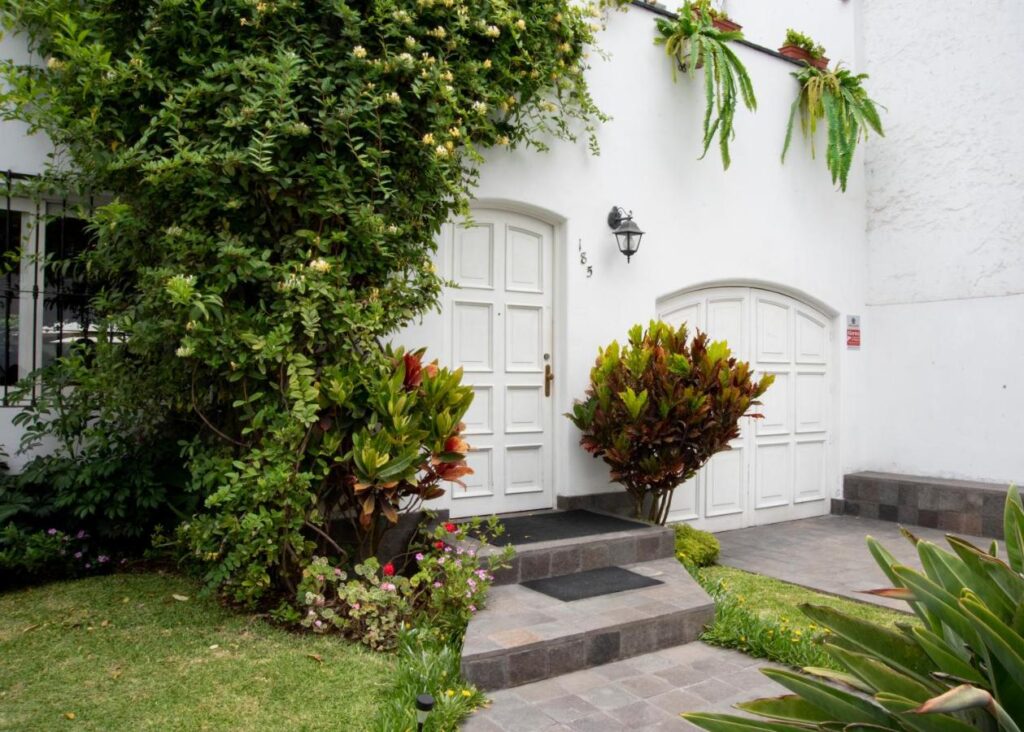 9 Unique Boutique Hotels in Lima 18 A white house with a white front door, arched garage door, and lush green plants evokes the charm of boutique hotels in Lima. Steps lead to the entrance, hanging ferns are above the garage, and house number 159 is by the door.