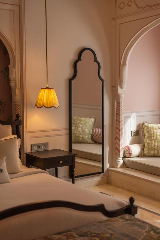 12 BEST Boutique Hotels in Jaipur 48 A cozy bedroom inspired by boutique hotels in Jaipur, with a cushioned window seat, a tall ornate mirror, wooden side table, and hanging yellow lampshade—all bathed in warm, soft light and elegant, light-colored decor.