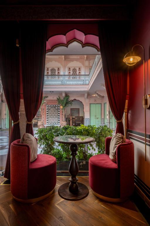 12 BEST Boutique Hotels in Jaipur 51 Two red velvet chairs with white pillows face each other at a small round table by tall windows, red curtains framing a lush courtyard—an inviting scene often found in boutique hotels in Jaipur’s ornate, historic buildings.