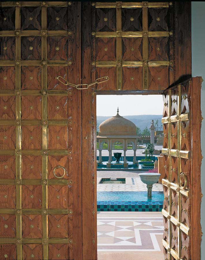12 BEST Boutique Hotels in Jaipur 44 A large ornate wooden door with brass detailing opens to reveal a courtyard reminiscent of boutique hotels in Jaipur, featuring a domed pavilion, fountains, and patterned tiles set against distant hills.