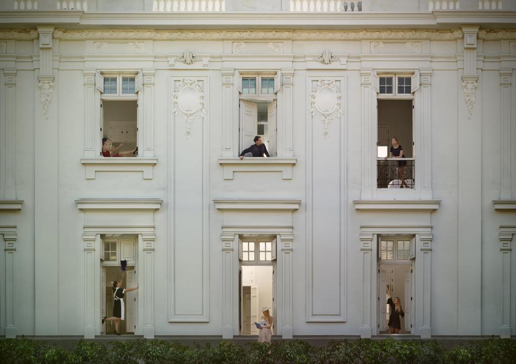 9 Unique Boutique Hotels in Lima 28 A white neoclassical building facade with six large windows, each revealing people engaged in different activities, such as reading, leaning out, or looking outside—just like the inviting charm of boutique hotels in Lima. Green shrubs line the bottom of the building.
