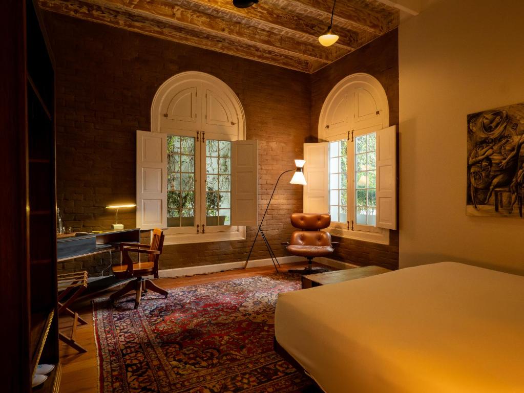 9 Unique Boutique Hotels in Lima 11 A cozy bedroom with warm lighting, inspired by boutique hotels in Lima, Peru—featuring a bed, patterned rug, wooden desk with a lamp, leather armchair, large arched windows with white shutters, and exposed wooden ceiling beams.