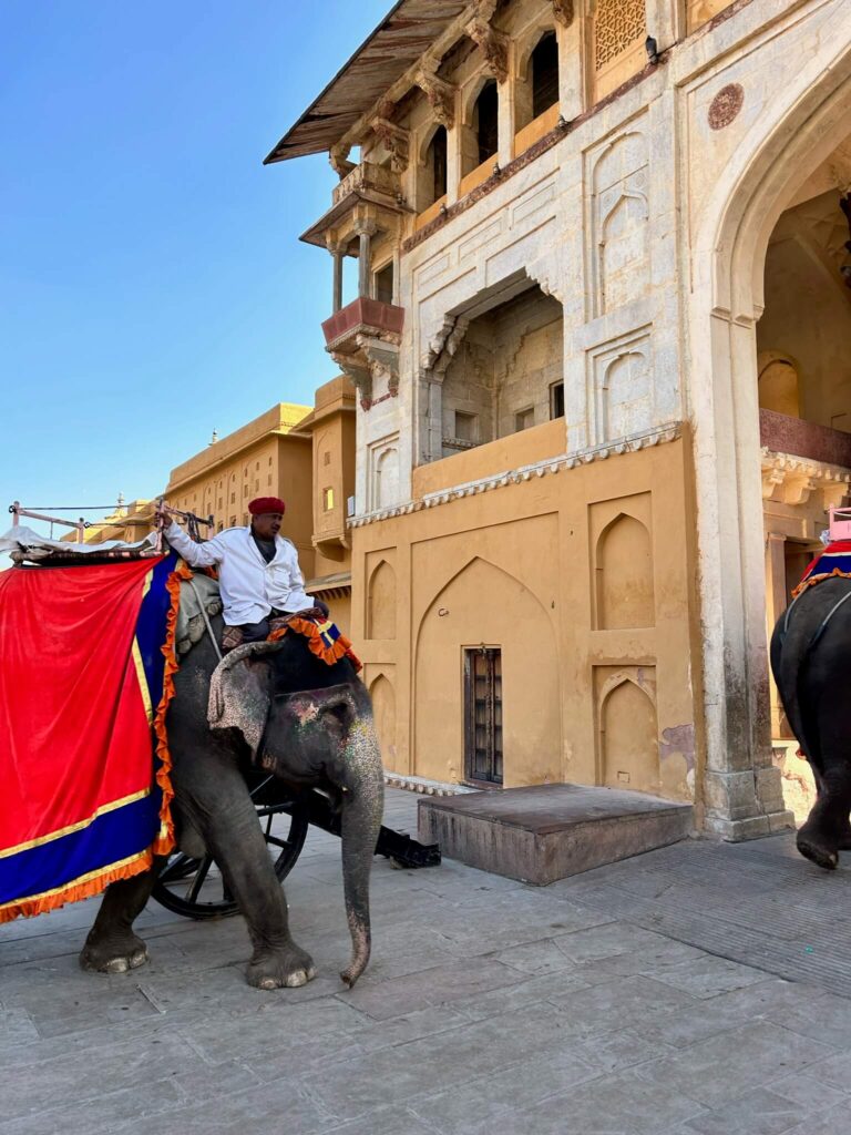 12 BEST Boutique Hotels in Jaipur 2 A man in traditional attire rides a decorated elephant near a historic yellow and white building with arches, reminiscent of the grandeur seen at boutique hotels in Jaipur. Another adorned elephant is partially visible on the right under the clear blue sky.