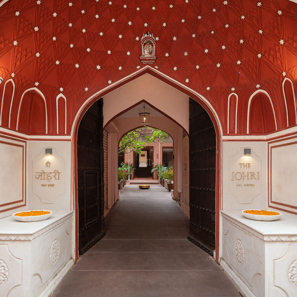 12 BEST Boutique Hotels in Jaipur 38 An ornate arched entrance with open wooden doors leads to a courtyard, echoing the charm of boutique hotels in Jaipur. The red and white archway is adorned with patterns and signs reading The Johri, with flower bowls placed on either side and inside.