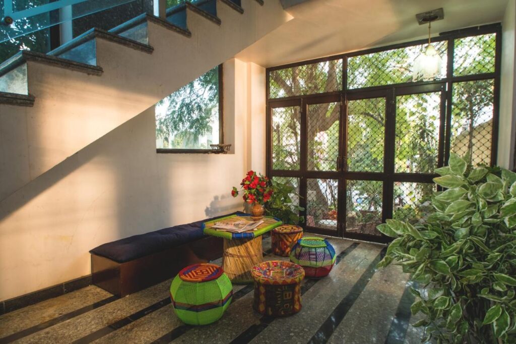 12 BEST Boutique Hotels in Jaipur 43 A cozy indoor seating area under a staircase, inspired by boutique hotels in Jaipur, with colorful poufs, a cushioned bench, potted plants, and large windows that flood the space with natural light.