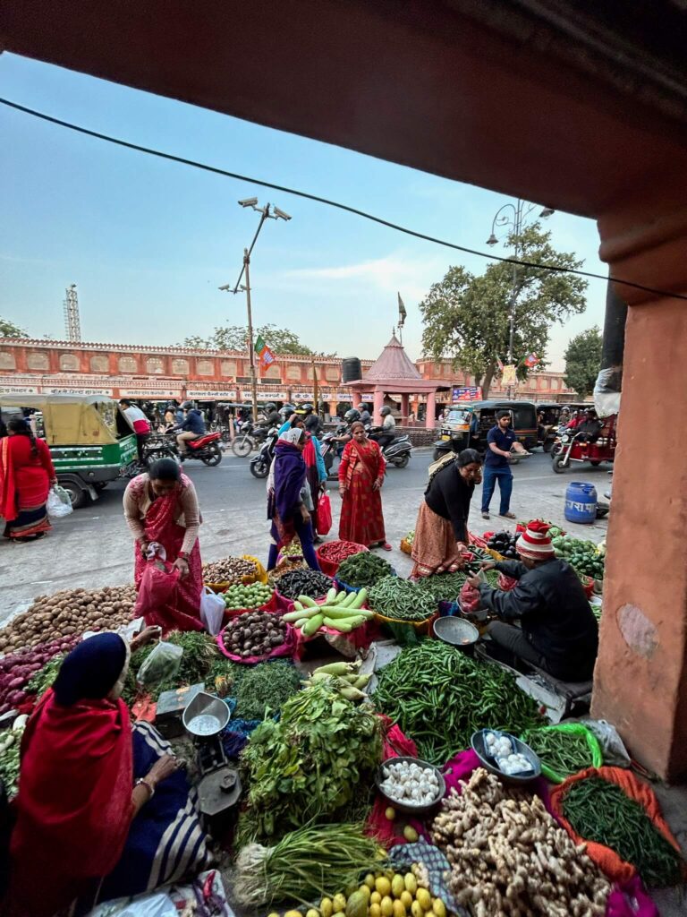 12 BEST Boutique Hotels in Jaipur 3 A busy outdoor market scene near boutique hotels in Jaipur, with vendors selling fresh vegetables on the pavement, shoppers browsing, and traffic including bikes and rickshaws passing by in the background under a clear sky.
