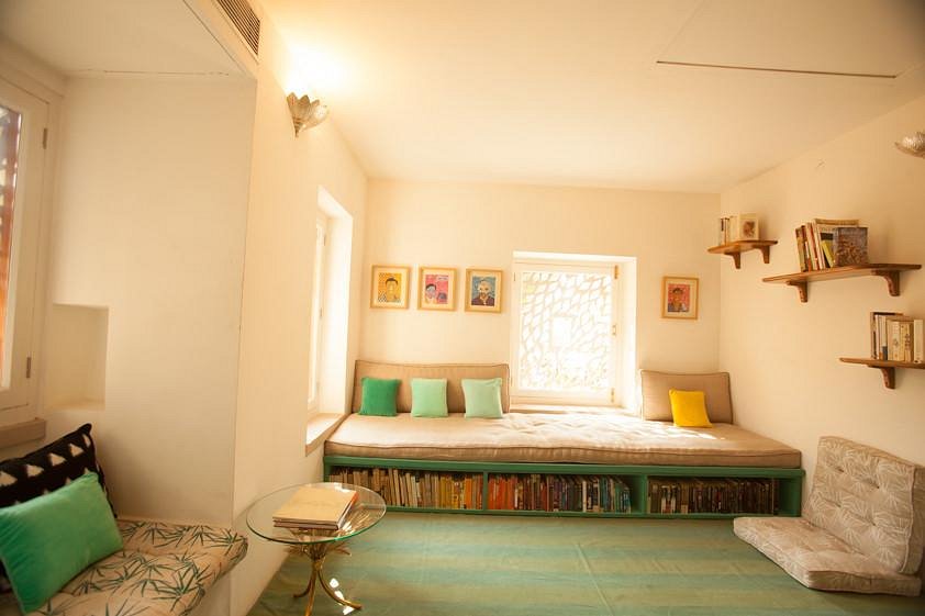 12 BEST Boutique Hotels in Jaipur 35 Bright, cozy reading nook with a large daybed, colorful cushions, bookshelves, and framed art—reminiscent of the inviting corners found in boutique hotels in Jaipur—set in a sunlit room with pale walls and a green rug.