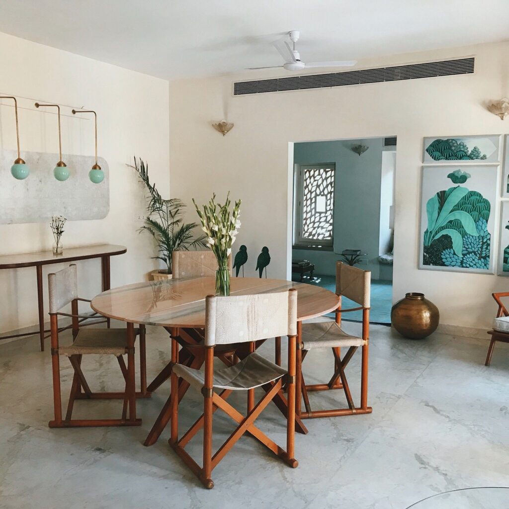 12 BEST Boutique Hotels in Jaipur 32 A modern dining room inspired by boutique hotels in Jaipur, featuring a round glass table, six wooden chairs with beige seats, green pendant lights, potted plants, and botanical wall art on a crisp white wall.