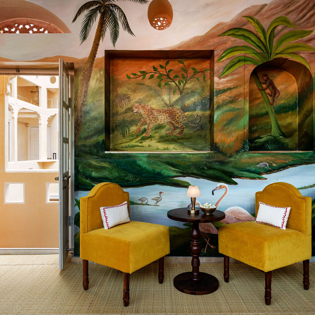 12 BEST Boutique Hotels in Jaipur 36 Two bright yellow chairs and a small round table sit in front of a vibrant jungle mural, reminiscent of the eclectic charm found in boutique hotels in Jaipur. An open door reveals an inviting outdoor area.
