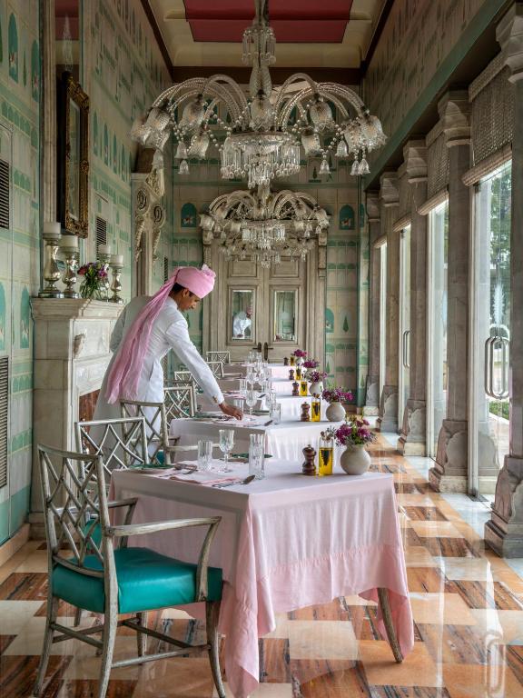 12 BEST Boutique Hotels in Jaipur 13 A waiter in a pink turban and sash sets a table in an elegant, sunlit dining room—reminiscent of the finest boutique hotels in Jaipur—with chandeliers, pastel decor, pink tablecloths, fresh flowers, and sparkling glassware adorning each table.