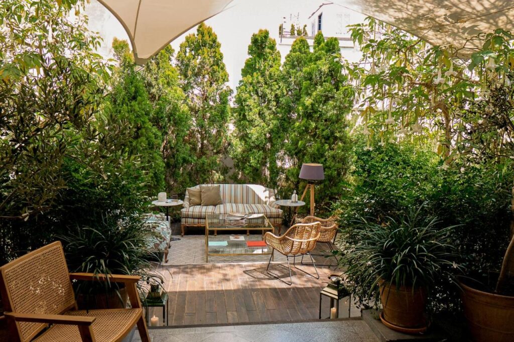 9 Unique Boutique Hotels in Lima 7 A cozy outdoor patio at one of Lima's boutique hotels features a sofa, wicker chairs, small tables, books, and a floor lamp, all surrounded by lush green plants and trees under a white canopy.