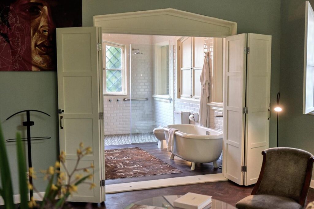 9 Unique Boutique Hotels in Lima 8 A stylish bathroom in the style of boutique hotels in Lima, with white double doors, a freestanding bathtub, brown rug, glass-enclosed shower, white subway tiles, large windows, and cozy decor. Part of an elegant room with green walls—perfect for Lima Peru inspiration.