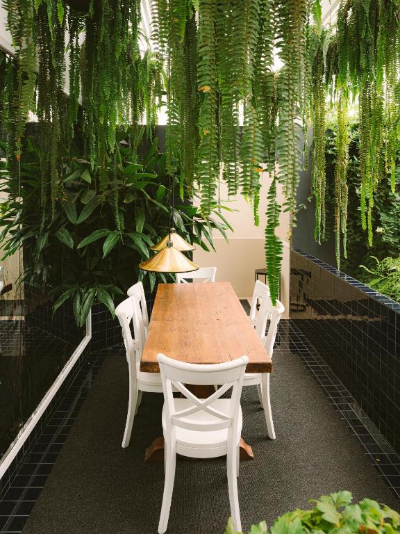 9 Unique Boutique Hotels in Lima 25 A wooden dining table with six white chairs sits beneath a hanging brass light, surrounded by lush green plants and ferns—evoking the stylish, plant-filled lounges of boutique hotels in Lima—on dark tiled floors.