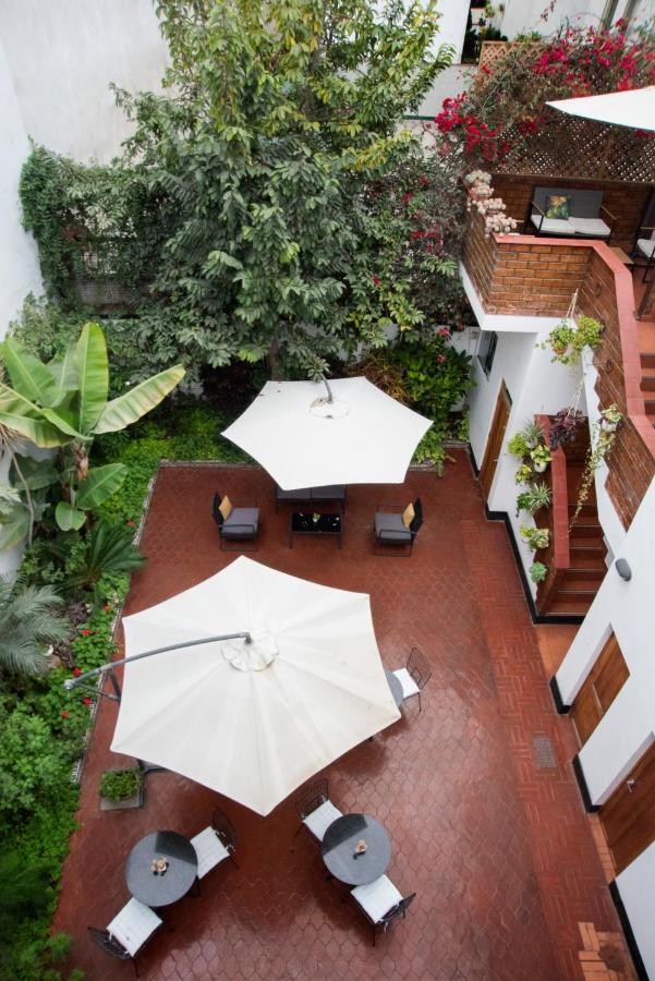 9 Unique Boutique Hotels in Lima 17 A view from above of a brick patio courtyard at one of the boutique hotels in Lima, featuring white umbrellas, round tables, chairs, lush green plants, and a staircase leading to a flower-adorned second level with potted plants.