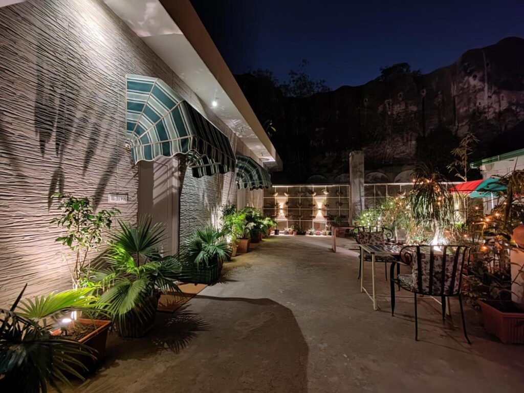12 BEST Boutique Hotels in Jaipur 42 A cozy outdoor patio at night, like those found in boutique hotels in Jaipur, features potted plants, decorative lighting, striped awnings, and cushioned metal chairs. Warm lights enhance the stone wall and rocky cliff visible in the background.