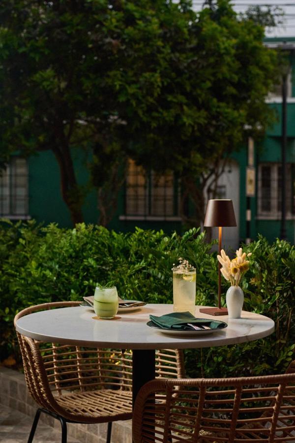 9 Unique Boutique Hotels in Lima 16 A round white table with two drinks, a green one and a tall yellow one, sits on a patio with wicker chairs—evoking the charming outdoor spaces of boutique hotels in Lima Peru, complete with flowers, a modern lamp, and lush green bushes and trees.