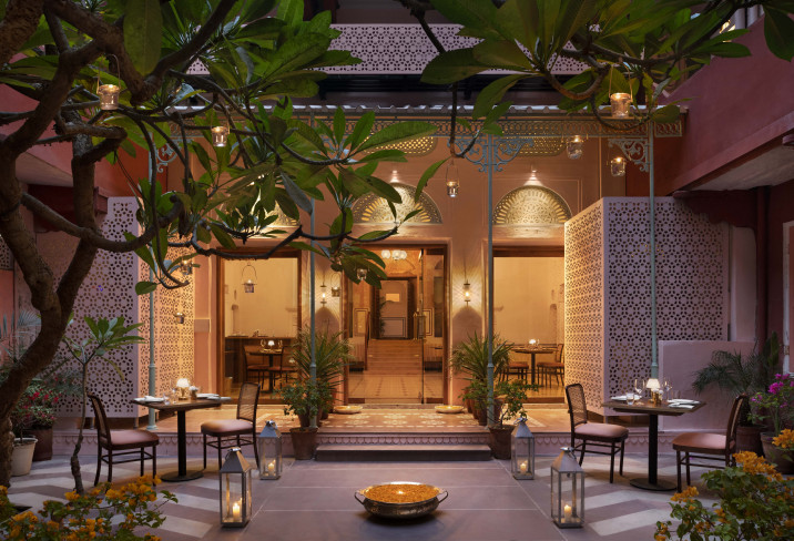 12 BEST Boutique Hotels in Jaipur 39 A softly lit outdoor restaurant patio at one of the boutique hotels in Jaipur features elegant tables and chairs, lanterns, lush green plants, and intricate decorative walls, creating a warm and inviting ambiance for dining.