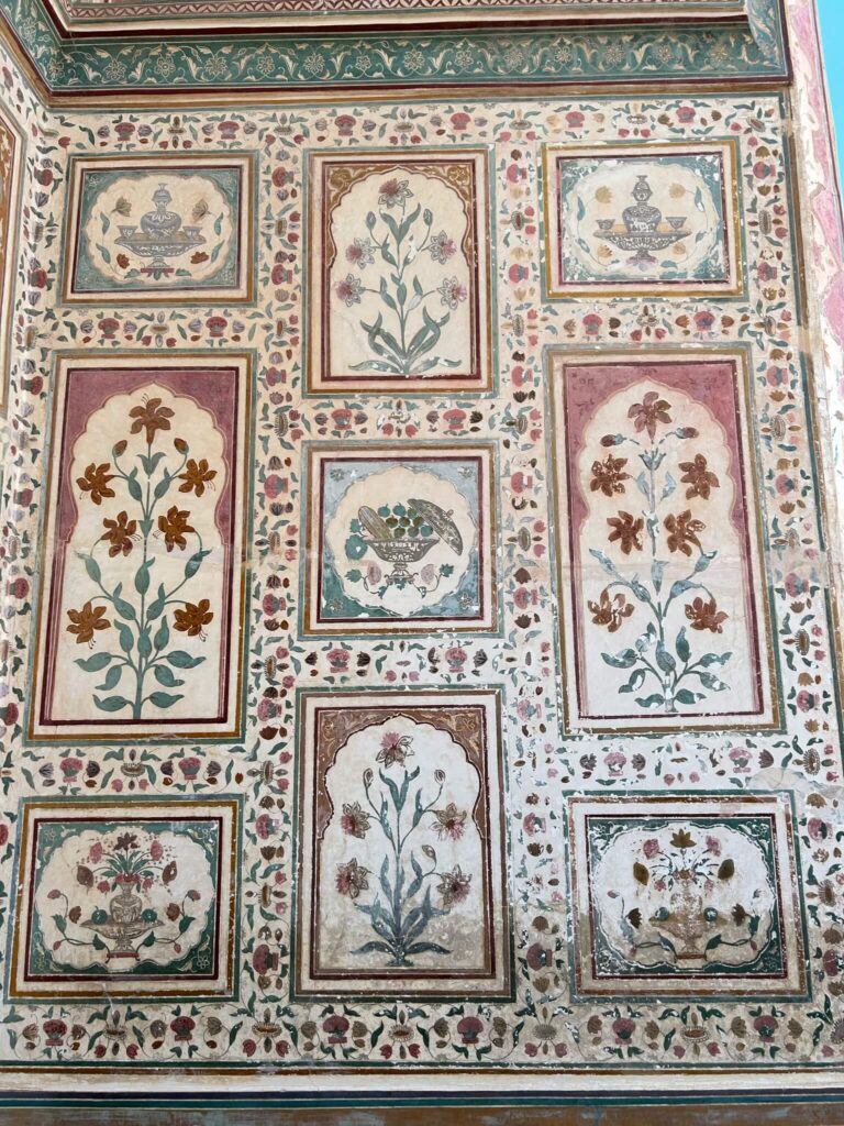 12 BEST Boutique Hotels in Jaipur 53 Ornate wall with rectangular and square panels featuring intricate floral designs and decorative borders in red, green, and beige tones—reminiscent of the elegant interiors found in boutique hotels in Jaipur. Each panel displays unique flower arrangements.