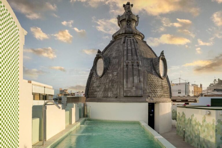 A rooftop swimming pool with green-and-white tile accents sits beside a large, dome-shaped structure with round windows and ornate details—an unforgettable scene among the boutique hotels in Valencia, set against a sunset sky with scattered clouds.