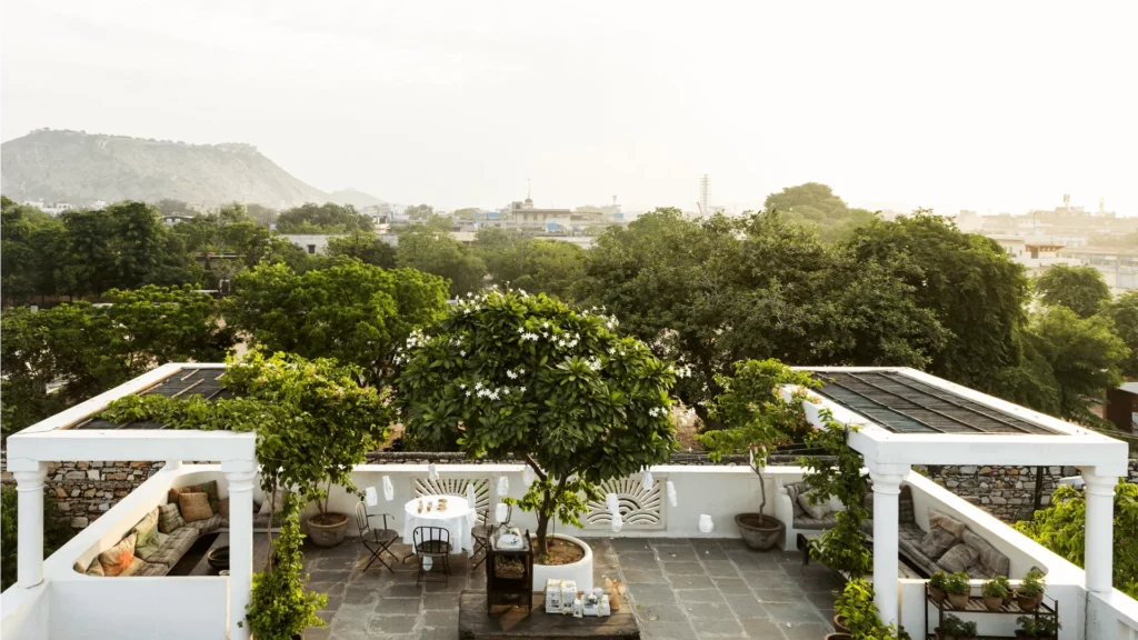 12 BEST Boutique Hotels in Jaipur 4 A rooftop terrace at one of the boutique hotels in Jaipur, featuring two shaded seating areas, a central table, lush potted plants, and surrounding greenery overlooking the cityscape and distant hills on a bright day.