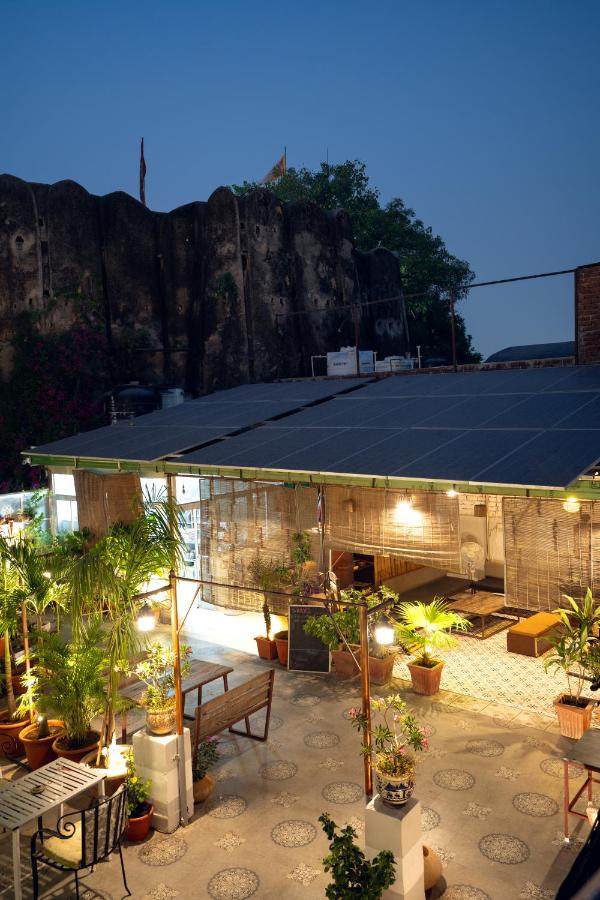 12 BEST Boutique Hotels in Jaipur 41 A cozy rooftop patio at dusk features potted plants, benches, string lights, and a covered seating area—much like the charming atmospheres found at boutique hotels in Jaipur—with an old stone wall and greenery in the background.