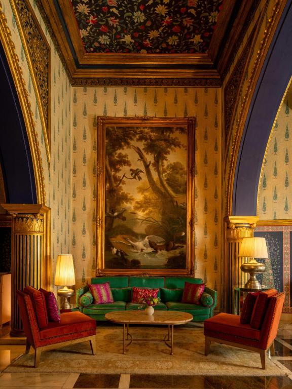 12 BEST Boutique Hotels in Jaipur 14 Luxurious lounge with a green velvet sofa, two red armchairs, a marble coffee table, and gold lamps—evoking the charm of boutique hotels in Jaipur. A large ornate painting and floral-patterned ceiling enhance the elegant, vintage decor.