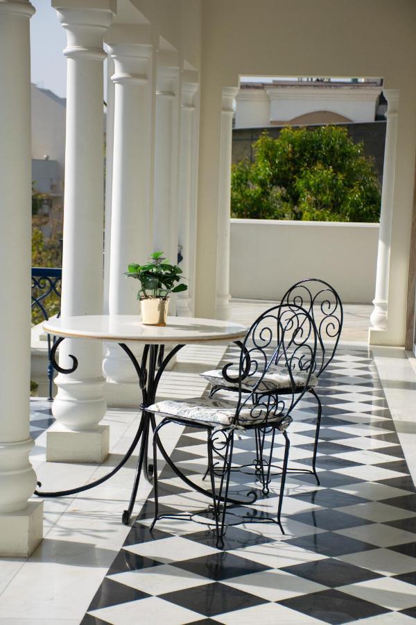 12 BEST Boutique Hotels in Jaipur 9 A sunlit veranda in the style of boutique hotels in Jaipur features a black-and-white checkered floor, white columns, and a small round table with two ornate metal chairs. A potted plant rests on the table, with trees visible in the background.