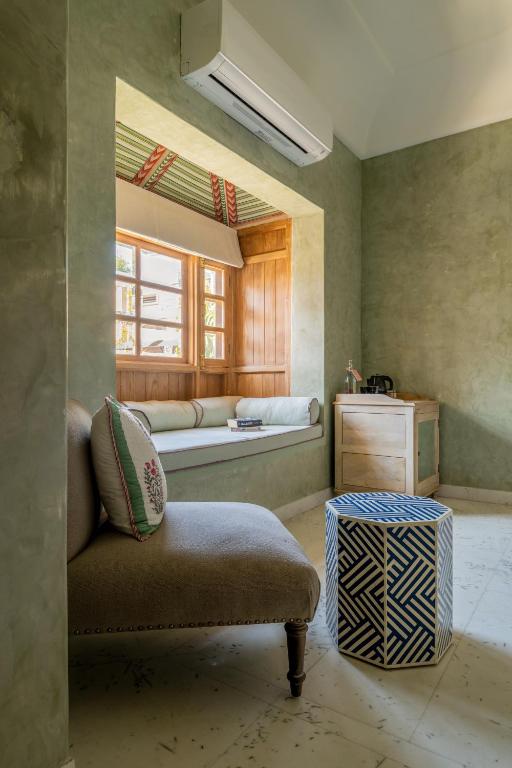 12 BEST Boutique Hotels in Jaipur 24 A cozy room in the style of boutique hotels in Jaipur, featuring a cushioned window seat, geometric blue-and-white ottoman, upholstered chair, small wooden cabinet with a kettle, and green textured walls under a split air conditioner.