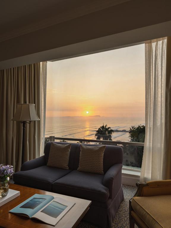 9 Unique Boutique Hotels in Lima 32 A cozy living room with a dark sofa, pillows, and a floor lamp sits by a large window overlooking the ocean at sunset—reminiscent of the serene lounges found in boutique hotels in Lima. Soft light fills the room, and a book is open on the coffee table.