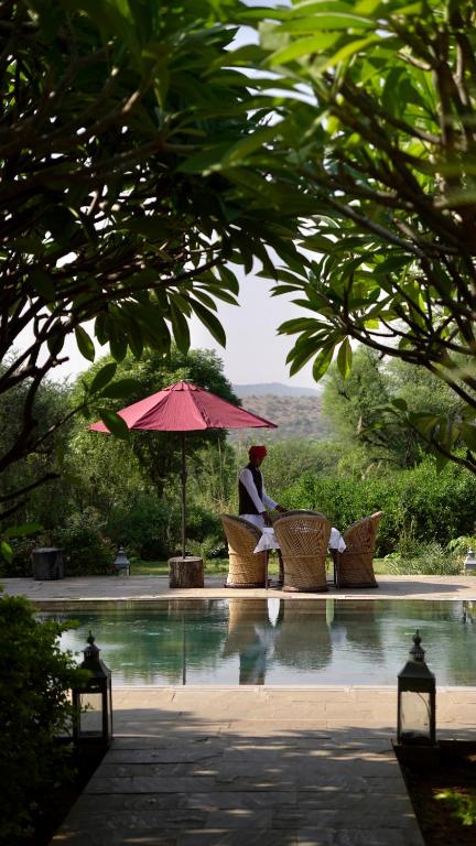 12 BEST Boutique Hotels in Jaipur 29 A man in traditional attire sits at a wicker table by a pool, shaded by a red umbrella—an inviting scene often found at boutique hotels in Jaipur, surrounded by lush greenery and framed by leafy trees.