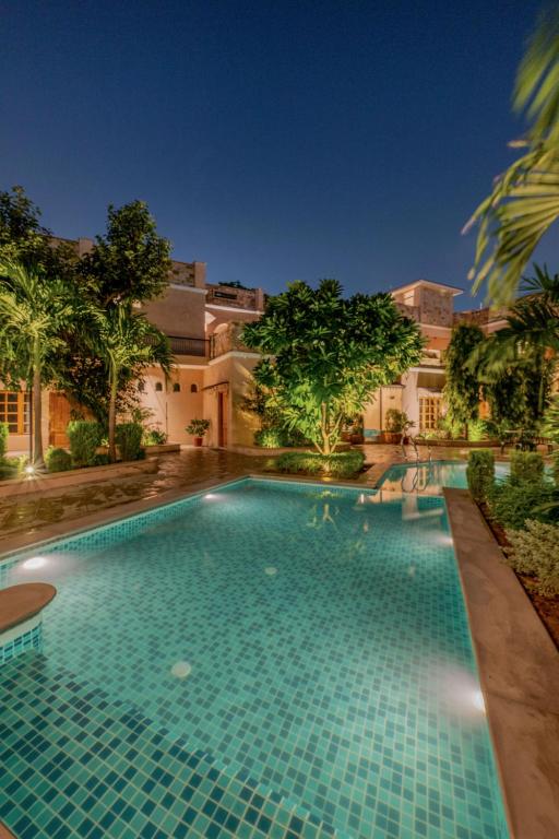 12 BEST Boutique Hotels in Jaipur 25 A well-lit swimming pool surrounded by lush greenery and palm trees, with a large house reminiscent of boutique hotels in Jaipur, all set under a clear night sky.