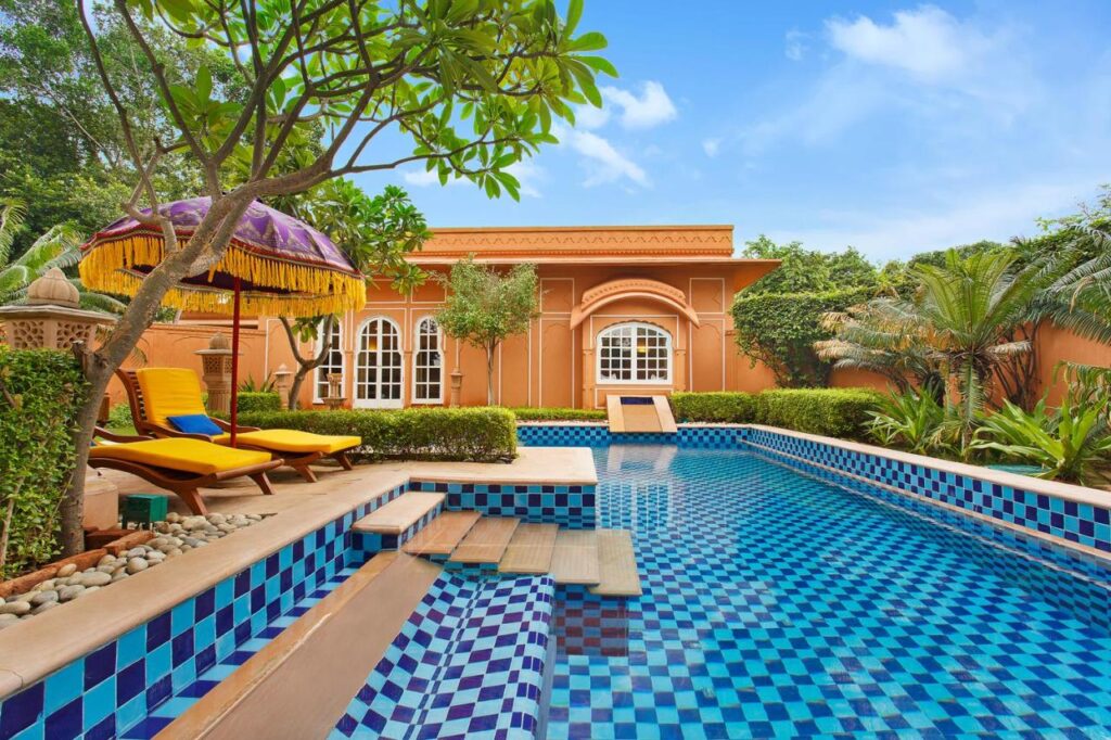 12 BEST Boutique Hotels in Jaipur 47 A vibrant, luxurious villa inspired by boutique hotels in Jaipur features orange walls and arched windows overlooking a mosaic-tiled swimming pool, surrounded by lush greenery, palm trees, and yellow lounge chairs under a decorative umbrella.