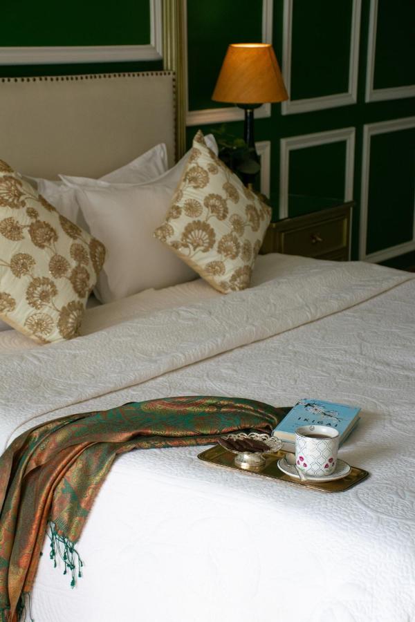 12 BEST Boutique Hotels in Jaipur 11 A neatly made bed with patterned pillows and a white bedspread, a green scarf, and a tray with coffee, snacks, and a blue book—evoking the relaxed elegance found in boutique hotels in Jaipur—beside a lit lamp and green-paneled wall.
