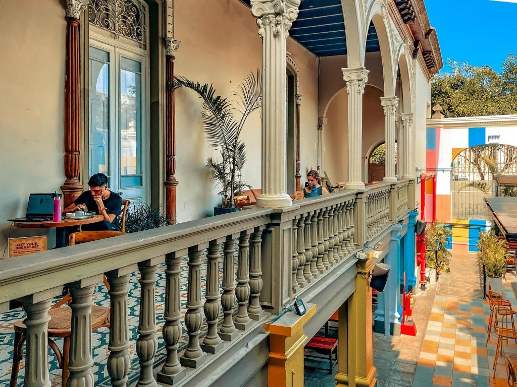 9 Unique Boutique Hotels in Lima 36 A man sits at a table with a laptop and coffee on a colorful, ornate balcony—much like those found in boutique hotels in Lima—overlooking a sunny courtyard filled with plants and vibrant walls.