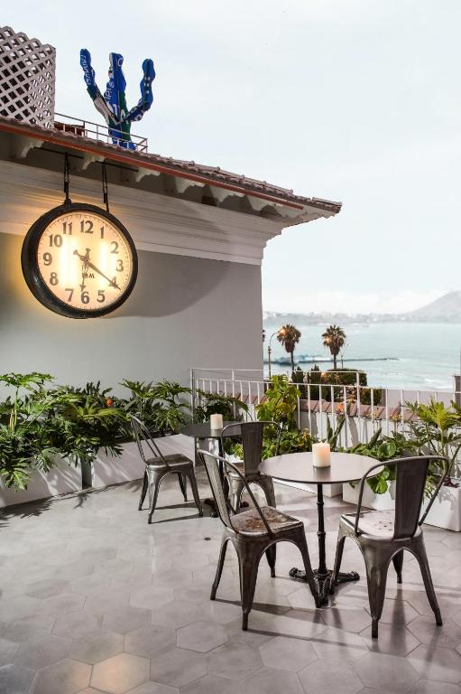 9 Unique Boutique Hotels in Lima 10 Outdoor patio with a round table, four metal chairs, potted plants, and candles. A large clock hangs on the wall, with a seaside view and palm trees—just like you'd find at boutique hotels in Lima Peru. Blue sculpture enhances the scene.